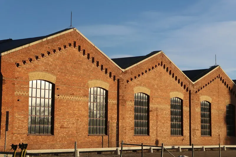 A red brick industrial building with a saw-tooth roof and tall arched windows